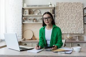 Portrait of a young beautiful woman working remotely at home in the kitchen using a laptop, with documents and a calculator. He is engaged in calculating the family budget. photo