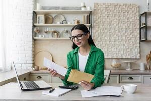 Smiling young woman working at home with laptop and documents. He opens the letter from the envelope he received. Got good news. photo