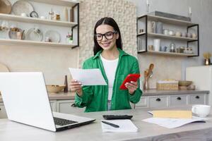 A young woman works and studies at home using a laptop. She is sitting in the kitchen, holding documents in her hands and using the phone. photo