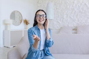 Beautiful young woman in headset sitting at home on sofa in front of camera and vlogging, recording blog, conducting webinar, growth and development trainings, pointing at camera, smiling. photo