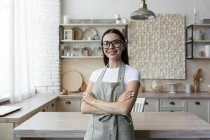 retrato de un joven blogger mujer en pie a hogar en el cocina en un delantal y mirando a el cámara. blogs acerca de hogar ciencias económicas y cocinando. foto