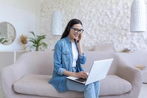 A young female student studies at home remotely on a laptop. He is sitting on the couch in a headset and jeans. Talks via call, zoom meeting, listens to online lectures and lessons. photo