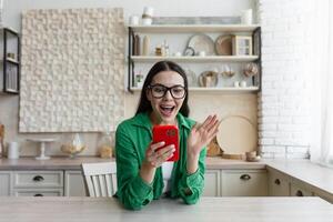 Game addict, game addiction. A young woman stands at home in the kitchen and plays online games, bets, lotteries on the phone. Screams with happiness, rejoices at the victory, financial reward. photo