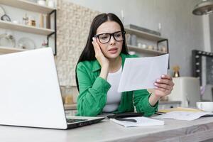 Sad young woman working from home with laptop and documents. Holds in hands and anxiously looks at bills, letter, message. Has problems with payment, debts. photo