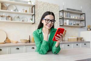 joven mujer en pie en cocina a hogar, propensión en mesa y utilizando móvil teléfono, charlando, comprobación social redes, enviando mensajes, mensajes de texto con amigos, novio. foto