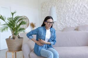 A young woman is sitting on the sofa at home, grimacing and holding her stomach. Suffers from severe abdominal pain, menstrual pain, pregnancy problems, miscarriage, poisoning, spasms. photo