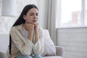 Young upset woman sitting on sofa at home, eyes closed, hands folded together near face. Suffers from depression, cries, thinks about solving problems. photo