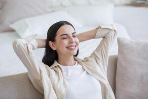 hermosa joven sonriente mujer relajante en sofá a hogar, sentado relajado con ojos cerrado y manos detrás cabeza. foto