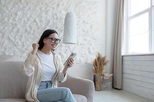 contento joven mujer sentado en sofá a hogar y utilizando móvil teléfono. él mira a el pantalla en sorpresa y alegría. foto