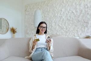 A young woman is sitting on the sofa at home and using the phone. Holds a credit card and makes remote purchases, rests, has fun. photo