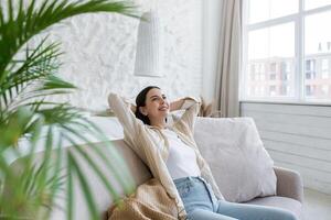 Young self-satisfied female student, business woman sitting on sofa at home and resting. Relaxed and dreamy, she threw her hands behind her head. photo