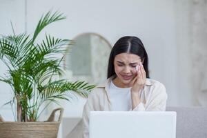 A young woman sits at home on the sofa with a notebook and cries, wipes her tears with a napkin. Received and read bad news, rummages on a call with fools, relatives, online psychologist. photo