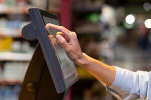 Close-up photo of a woman's hand in a supermarket inside using a self-service scale weighing vegetables and fruit in the store.