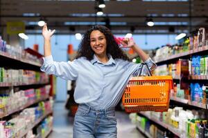 Portrait of a happy and smiling woman shopper in a supermarket, herpanic woman shopping and looking at the camera happily chooses products inside the store among the shelves with a shopping basket. photo