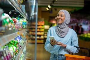 joven contento mujer comprador con teléfono elige productos en grande tienda de comestibles almacenar, musulmán mujer en hijab usos en línea compras lista, usos aplicación en teléfono inteligente foto