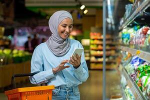 joven contento mujer comprador con teléfono elige productos en grande tienda de comestibles almacenar, musulmán mujer en hijab usos en línea compras lista, usos aplicación en teléfono inteligente foto