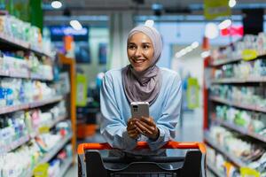 Shopping. A young beautiful Muslim woman in a hijab walks and chooses between the shelves with goods in a supermarket with a trolley. He is holding a phone with a list, application, online payment. photo