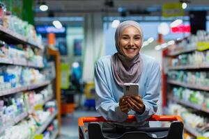 retrato de un comprador dentro un grande supermercado almacenar, un musulmán mujer en un hijab con un compras carro usos un teléfono inteligente aplicación, elige productos con descuentos y sonrisas foto
