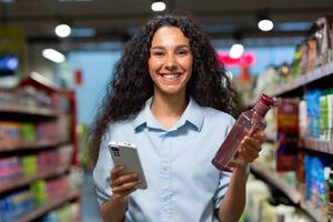 Portrait of a female buyer in a supermarket, a Hispanic woman chooses shampoo for hair and shower gel, reads reviews on the Internet, smiles and uses an application on her phone. photo