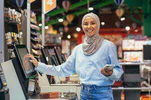 autoservicio en un supermercado. un joven árabe mujer en un hijab es en pie cerca el electrónico efectivo registros y es pago para el bienes en el Tienda en línea mediante un solicitud en su teléfono. foto