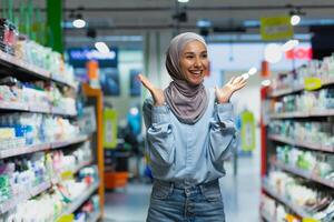 Shopping. A young Muslim woman in a hijab walks between the shelves with goods in a supermarket, chooses, spreads her hands, is surprised by the large selection, is satisfied with the variety. photo