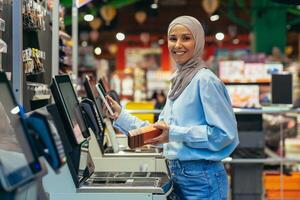 autoservicio en un supermercado. un joven árabe mujer en un hijab es en pie cerca el electrónico efectivo registros y es pago para el bienes en el Tienda en línea mediante un solicitud en su teléfono. foto