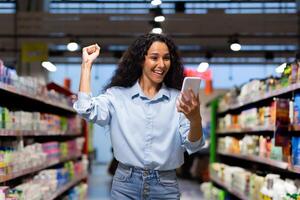 alegre joven hembra comprador levanta su puño en victoria mientras mirando a su teléfono en un tienda de comestibles Tienda pasillo. foto