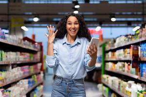 alegre joven mujer ondulación a teléfono mientras chateando en un almacenar, expresando felicidad y conectividad en medio de compras. foto