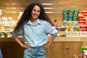 A cheerful woman with curly hair stands hands on hips in a cozy pottery and home decor store, exuding confidence and positivity. photo