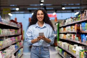 sonriente joven hembra comprador utilizando un móvil teléfono en el tienda de comestibles almacenar, en medio de el vistoso estantería lleno de productos foto