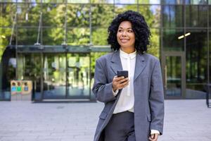 exitoso africano americano negocio mujer caminando en el ciudad desde fuera de oficina edificio, financiero trabajador sonriente contentamente, mujer participación teléfono inteligente utilizando aplicación, hojeada social redes foto