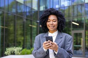 joven exitoso satisfecho negocio mujer caminando con teléfono en manos, africano americano mujer en negocio traje con Rizado pelo participación teléfono inteligente, sonriente hojeada social medios de comunicación. foto