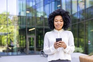 Young beautiful African-American woman walking street outside office building, businesswoman holding a phone i hands, smiling contentedly, browsinginternet and using an application on a smartphone. photo