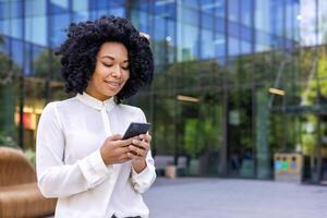 Young beautiful African-American woman walking street outside office building, businesswoman holding a phone i hands, smiling contentedly, browsinginternet and using an application on a smartphone. photo