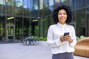 Young beautiful African-American woman walking street outside office building, businesswoman holding a phone i hands, smiling contentedly, browsing internet and using an application on a smartphone. photo