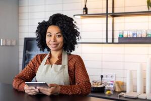 retrato camarera de un pequeño negocio propietario de un cafetería, mujer en delantal sonrisas y mira a cámara con un tableta computadora en su manos, un empleado de un café de un restaurante cerca un café encimera. foto