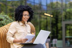 Smiling young African American female student sitting on a bench on a university campus studying online using a laptop. Writes in a notebook and listens to a lecture. photo