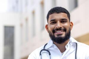 Close-up portrait of a young Indian male doctor standing outside a clinical center in a white coat and smiling at the camera. photo
