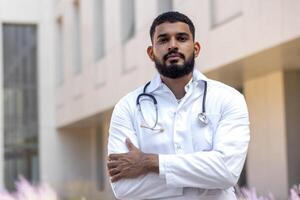 Serious young muslim male doctor standing outside clinic outside wearing a robe and holding a stethoscope and crossing his arms looking confidently at the camera. photo