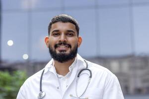 Close-up portrait of a young Muslim male doctor, student standing outside in a white coat and holding a stethoscope and looking confidently and smiling at the camera. photo