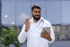 un sonriente joven masculino indio médico soportes en el calle cerca el hospital en un blanco Saco y comunica vía llamada con pacientes, consulta remotamente en línea, saluda y olas a el cámara. foto