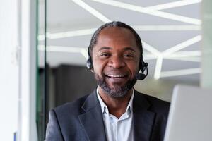 Close-up portrait of a senior smiling African-American man working in an office using a laptop in a headset and smiling at the camera. photo