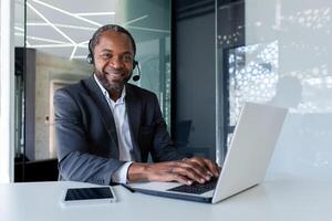Portrait of african american businessman inside office at workplace, mature man using headset phone and laptop for remote consultation with clients, smiling and looking at camera. photo