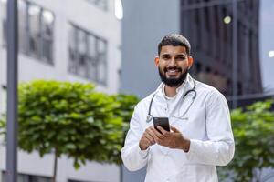 Portrait of a young Muslim male doctor standing smiling near the clinic and holding a phone in his hands. Looking and smiling at the camera. photo