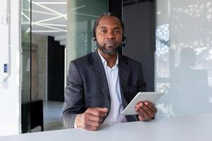 Portrait of a serious senior african man who works in the office in a headset, sits at the table, looks seriously into the camera, uses a tablet. photo