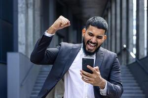 A happy young man in a business suit stands near an office building and looks at the phone, rejoices at success and received news, shows a victory gesture with his hand. photo