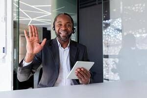 Help desk worker inside office at workplace, man smiling and looking at camera, waving greeting gesture, talking remotely, using headset phone and tablet for calling and consulting customer support photo