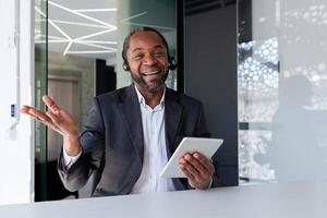 Portrait of a young African-American man working in the office, sitting at a desk in a headset, holding a tablet, smiling and gesturing to the camera. photo