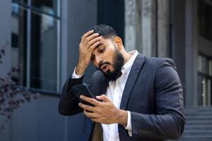 trastornado musulmán masculino empresario en pie cerca oficina y participación cabeza, conmocionado leyendo noticias, mensaje recibió. foto