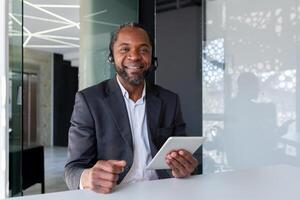 Portrait of support worker inside office at workplace, man smiling and looking at camera, using headset phone and tablet for communication and consulting customer support. photo
